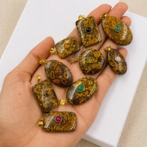May include: A collection of eight polished brown and yellow patterned jasper pendants in various shapes, including rectangular, oval, and circular. Each pendant features a small, smooth, colourful cabochon stone set in a gold-toned metal bail.