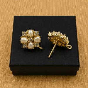 May include: Two gold-colored stud earrings with a floral design. Each earring features a cluster of white pearls and clear crystals. The earrings are displayed on a black box against a tan background.