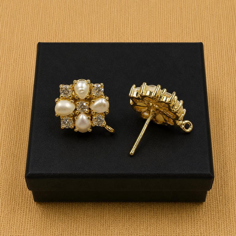 May include: Two gold-colored stud earrings with a floral design. Each earring features a cluster of white pearls and clear crystals. The earrings are displayed on a black box against a tan background.