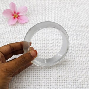 May include: A clear, circular bangle bracelet. The transparent bracelet is held by a hand, with a pink flower in the background. The bangle is made of a smooth, possibly acrylic material.