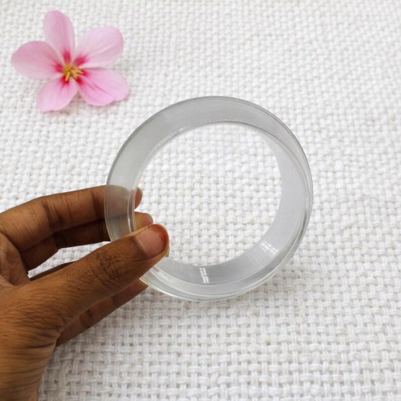 May include: A clear, circular bangle bracelet. The transparent bracelet is held by a hand, with a pink flower in the background. The bangle is made of a smooth, possibly acrylic material.