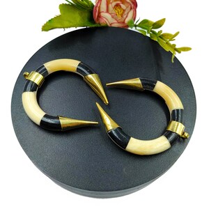 Peut inclure: Une paire de pendentifs incurv&eacute;s en forme de croissant avec des segments altern&eacute;s de couleur cr&egrave;me et noire. Chaque extr&eacute;mit&eacute; des pendentifs est coiff&eacute;e d'une pointe en m&eacute;tal dor&eacute; poli, et une extr&eacute;mit&eacute; comporte une petite boucle m&eacute;tallique.