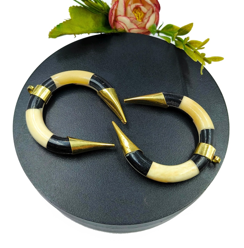 Peut inclure: Une paire de pendentifs incurv&eacute;s en forme de croissant avec des segments altern&eacute;s de couleur cr&egrave;me et noire. Chaque extr&eacute;mit&eacute; des pendentifs est coiff&eacute;e d'une pointe en m&eacute;tal dor&eacute; poli, et une extr&eacute;mit&eacute; comporte une petite boucle m&eacute;tallique.