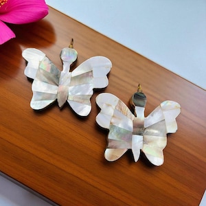 May include: Two butterfly-shaped pendants with iridescent mother-of-pearl inlay. The wings are segmented with a mix of white, cream, and iridescent pieces. Each pendant has a small gold-colored loop at the top for hanging.