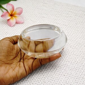 May include: A clear, round glass bowl with a thick base. The bowl is held in a hand, with a pink and yellow flower in the background. The bowl is likely used for decorative purposes.