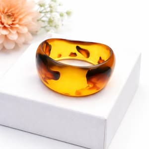 May include: A chunky, translucent bangle bracelet with a tortoiseshell pattern. The bracelet has amber and dark brown tones, creating a marbled effect. The jewellery is displayed on a white surface, highlighting its design.