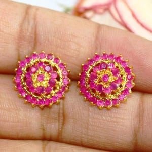 May include: A pair of gold earrings with pink gemstones set in a circular design. The earrings are small and delicate.