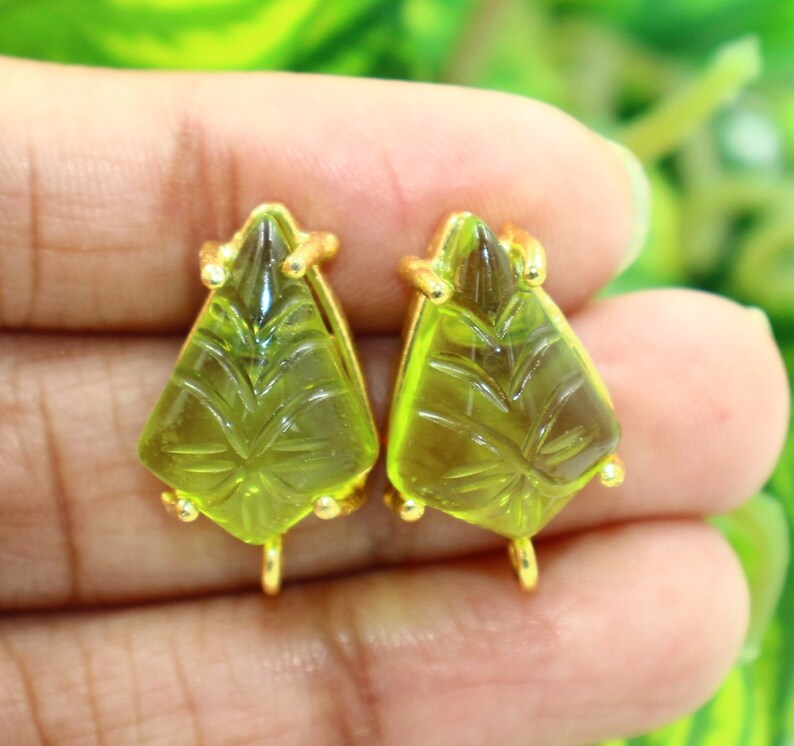 May include: A pair of gold-tone earrings with green glass teardrop-shaped stones. The stones have a carved design and are set in a gold-tone metal.