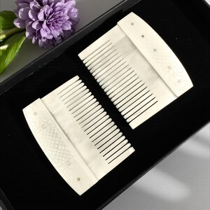 May include: Two ivory-colored combs with fine teeth, displayed in a black box. The combs have decorative geometric patterns and small, inlaid accents. A purple flower and green leaves are visible in the background.