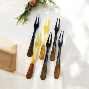 May include: A collection of dessert forks with a two-tone design. The forks have black and yellow tines with brown and yellow handles. The forks are arranged on a white surface with a floral arrangement in the background.