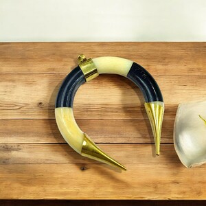 Peut inclure: Un objet incurv&eacute; en forme de croissant compos&eacute; de segments altern&eacute;s de mati&egrave;re lisse couleur cr&egrave;me et de mati&egrave;re noire polie. La pi&egrave;ce est agr&eacute;ment&eacute;e d'embouts en m&eacute;tal dor&eacute; poli aux deux extr&eacute;mit&eacute;s et d'une boucle centrale en m&eacute;tal dor&eacute;.