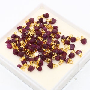 Puede incluir: Una colección de pequeños colgantes de rubí en bruto con anillas de salto chapadas en oro. Los rubíes son de color rojo intenso y tienen una textura rugosa y natural. Los colgantes son perfectos para añadir un toque de elegancia a los proyectos de joyería.