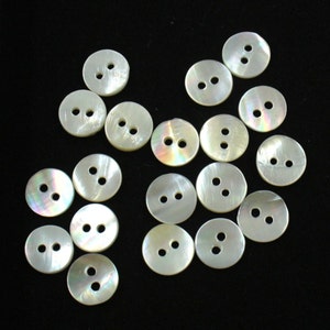 May include: A collection of 17 white iridescent buttons with two holes each. The buttons are arranged in a random pattern on a black background.
