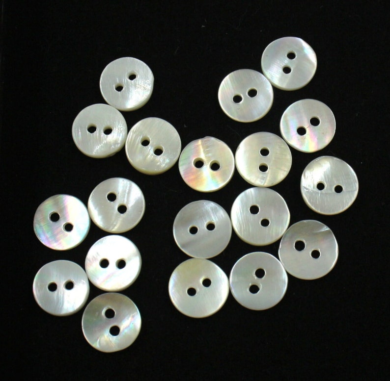 May include: A collection of 17 white iridescent buttons with two holes each. The buttons are arranged in a random pattern on a black background.