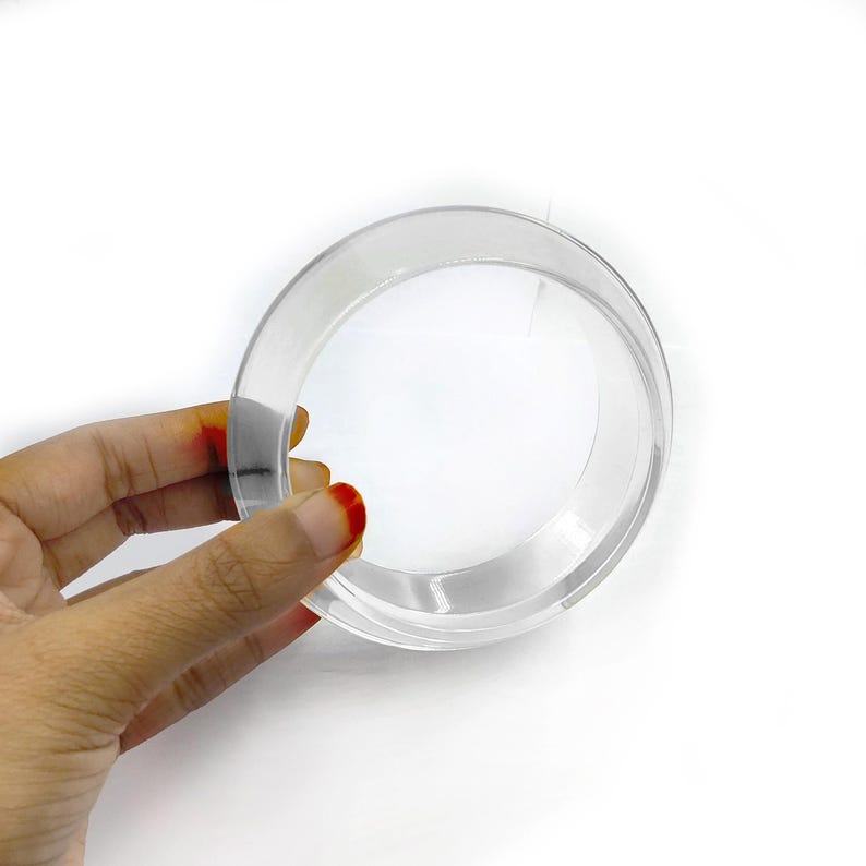 May include: A clear, circular, acrylic object with a flat base and a rounded rim. The object is held by a hand with red nail polish. The background is plain white.