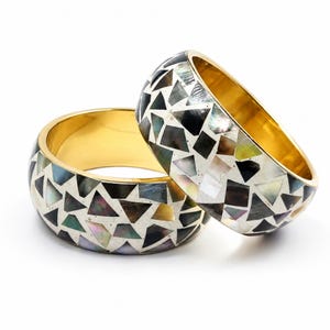 May include: Two wide bangle bracelets featuring a mosaic pattern of iridescent black, gray, and white shell fragments set in a silver-toned base with a smooth, polished gold-toned interior lining. The geometric inlay creates a reflective surface.
