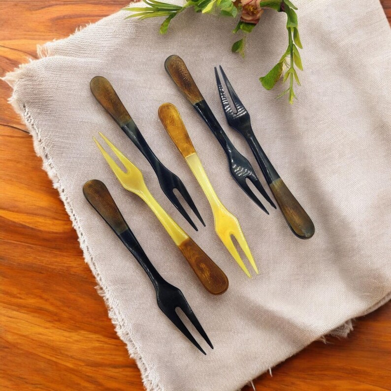 May include: A collection of six small forks with a two-pronged design. The forks have dark brown handles and black or yellow tines. They are arranged on a beige linen napkin, with a wooden table in the background. Decorative kitchenware.