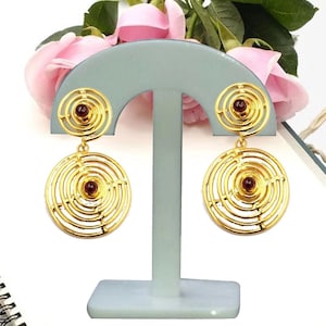 May include: Gold tone dangle earrings with a circular design and a red gemstone in the center. The earrings are displayed on a white stand.