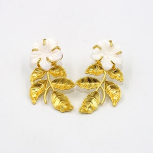 May include: A pair of floral earrings. Each earring features a white flower at the top, with a gold-colored stem and leaves. The earrings are set against a white background.