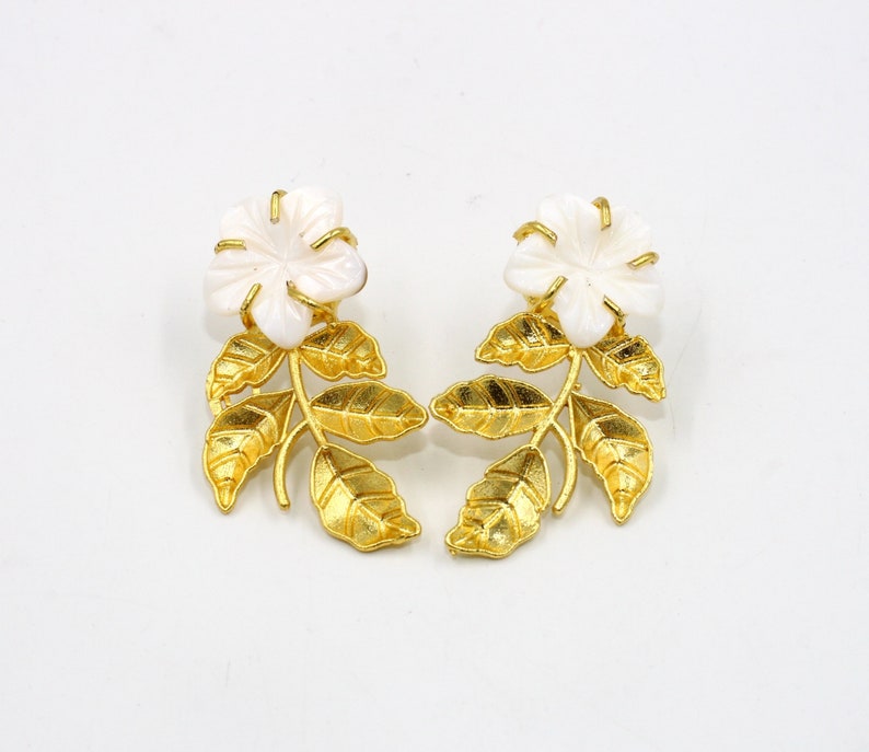 May include: A pair of floral earrings. Each earring features a white flower at the top, with a gold-colored stem and leaves. The earrings are set against a white background.