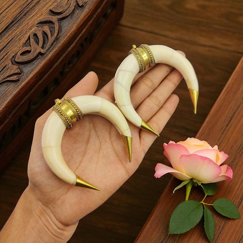 May include: Two ivory-coloured, curved earrings with gold-coloured metal accents. The earrings have a horn-like shape with a gold-coloured tip and a decorative band at the top. A pink and yellow rose is in the background.