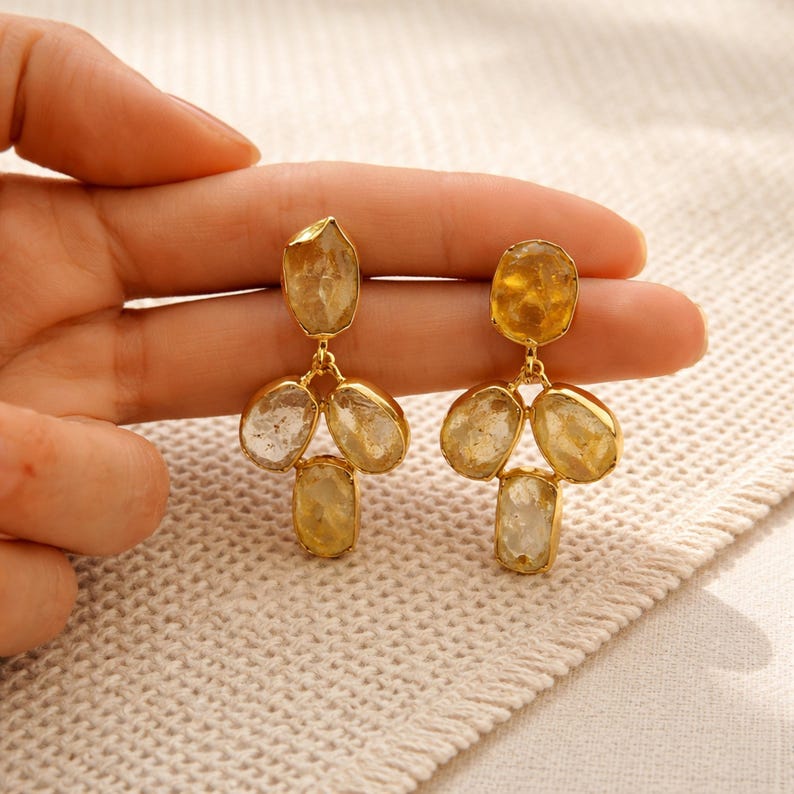 May include: Gold-toned earrings featuring three dangling, irregularly shaped, light yellow gemstones. The earrings have a post-back closure and are displayed on a cream-colored surface. The gemstones have a translucent quality, and the earrings are likely for pierced ears.