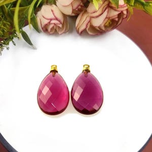 May include: Two faceted, teardrop-shaped pendants in a deep magenta hue, each topped with a small gold-colored cap. The pendants are displayed on a white surface, with floral accents in the background.