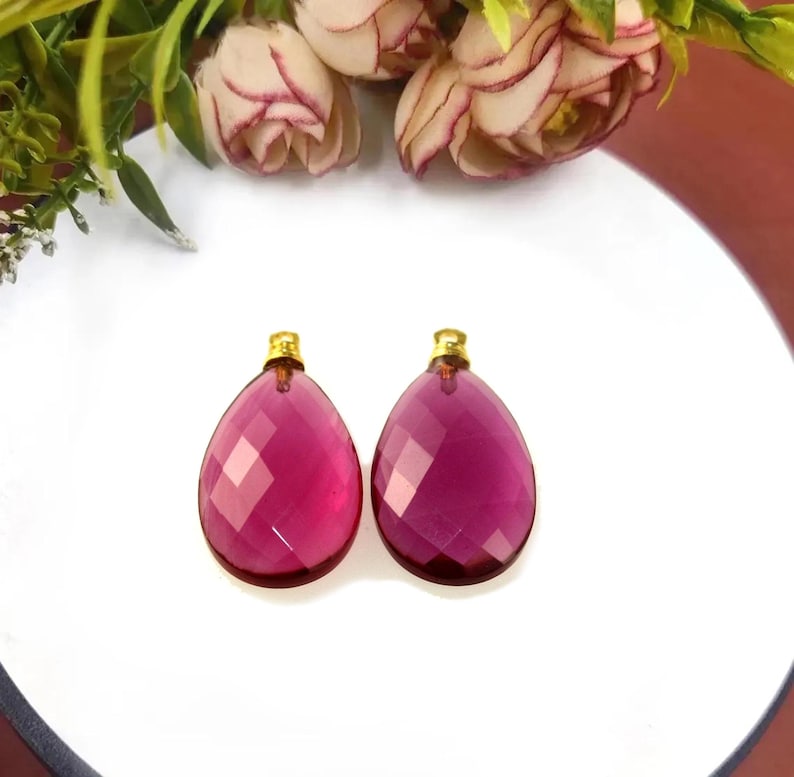 May include: Two faceted, teardrop-shaped pendants in a deep magenta hue, each topped with a small gold-colored cap. The pendants are displayed on a white surface, with floral accents in the background.