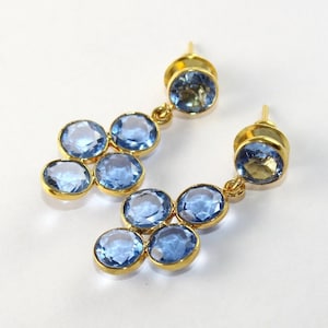 May include: A pair of gold earrings with blue gemstone accents. The earrings feature a cluster of five round gemstones, each set in a gold setting. The earrings are dangle style, with a single larger gemstone at the bottom.