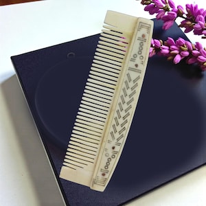 May include: Ivory-colored comb with fine teeth and decorative etched patterns. The comb features a series of small circles and diagonal lines along the handle. The comb is resting on a dark blue surface, with a sprig of purple flowers in the background.