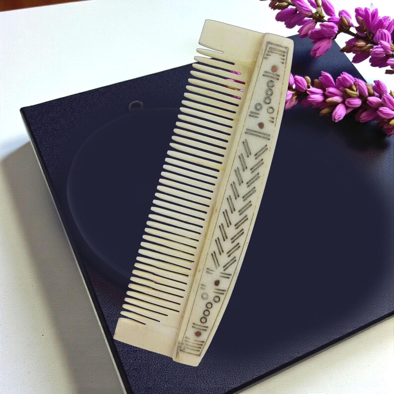 May include: Ivory-colored comb with fine teeth and decorative etched patterns. The comb features a series of small circles and diagonal lines along the handle. The comb is resting on a dark blue surface, with a sprig of purple flowers in the background.