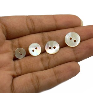 May include: Four white mother of pearl buttons with two holes each. The buttons are arranged in a row on a hand.