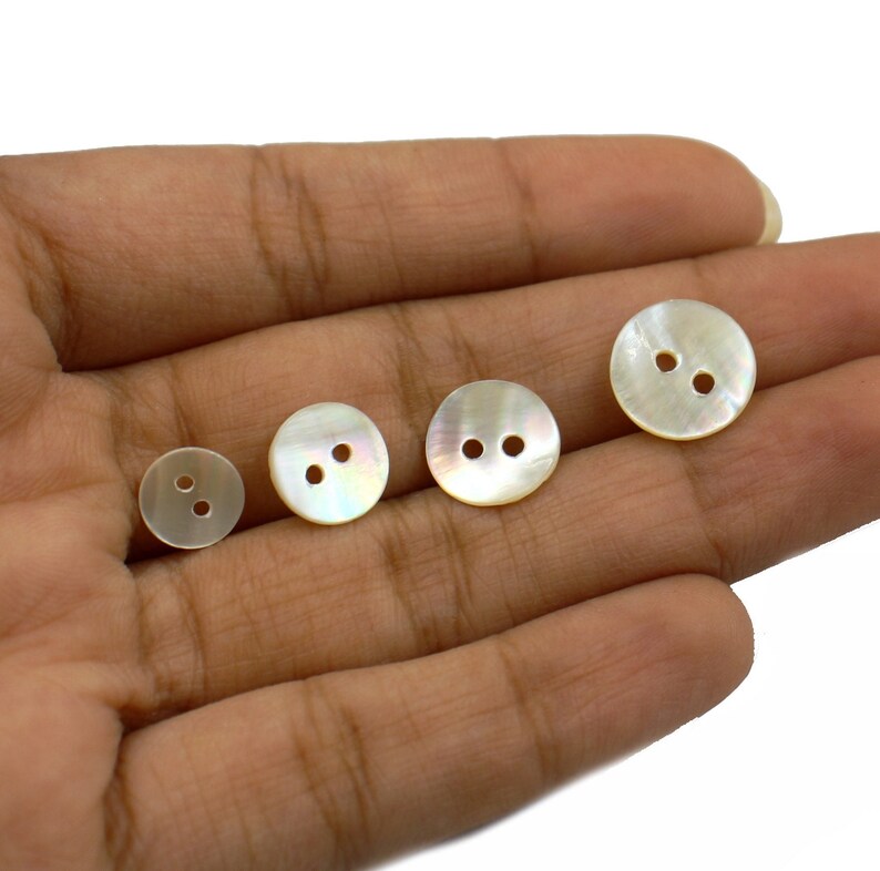 May include: Four white mother of pearl buttons with two holes each. The buttons are arranged in a row on a hand.