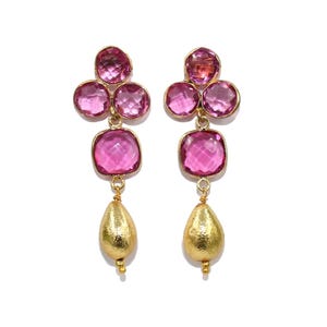 Puede incluir: Un par de pendientes de oro con gotas de piedras preciosas rosas. Los pendientes presentan un grupo de cuatro piedras preciosas rosas en la parte superior, seguido de una piedra preciosa rosa cuadrada más grande y una perla dorada en forma de lágrima.