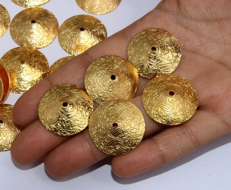 May include: A collection of gold-toned metal embellishments with a decorative floral pattern. The embellishments are round and have a small hole in the center.