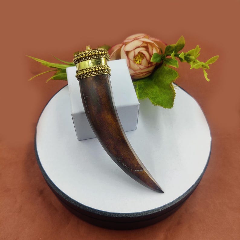 May include: A dark brown wooden pendant shaped like a tusk with a gold metal top. The pendant is on a white circular display stand.