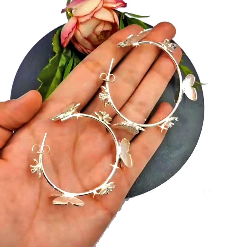 Puede incluir: Pendientes de aro de plata con abalorios de mariposa. Los pendientes son grandes y tienen un dise&ntilde;o delicado.