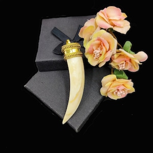 May include: A white and gold pendant shaped like a tusk with a gold band around the top. The pendant is sitting on a black box with a bouquet of pink flowers.