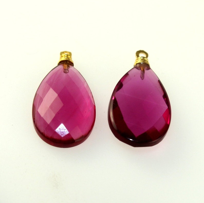 May include: Two faceted, teardrop-shaped pendants. The pendants are a deep magenta color with gold-colored metal caps. The facets create a sparkling effect. These pendants are suitable for jewelry making.