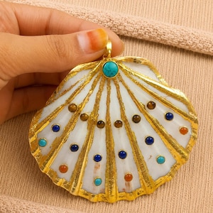 May include: A large, ornate pendant in the shape of a seashell. The pendant features a white base with gold-colored accents and is adorned with various colorful gemstones, including turquoise, brown, blue, and orange. The pendant is held by a hand.