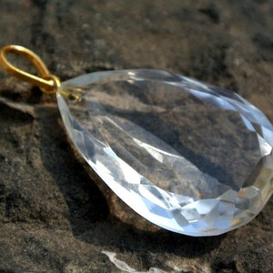 May include: A clear, faceted crystal pendant with a gold-colored bail. The teardrop-shaped gemstone is transparent, reflecting light. The pendant is set against a dark, textured background.