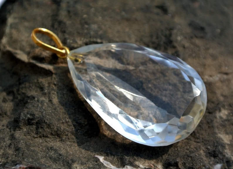 May include: A clear, faceted crystal pendant with a gold-colored bail. The teardrop-shaped gemstone is transparent, reflecting light. The pendant is set against a dark, textured background.