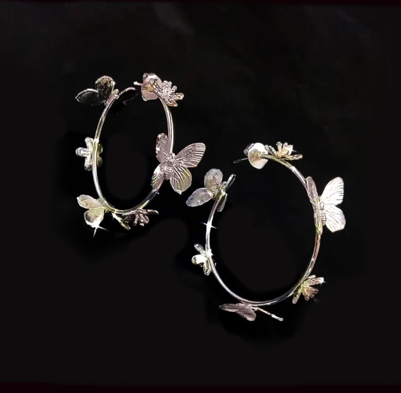 Puede incluir: Un par de pendientes de aro plateados con detalles de mariposa y flores. Los pendientes est&aacute;n hechos de metal y tienen un dise&ntilde;o delicado y femenino.
