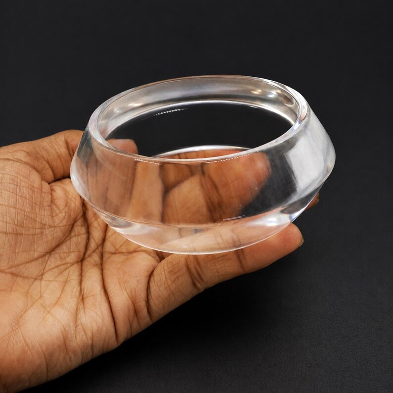 May include: A clear, transparent bangle bracelet with a smooth, polished surface. The circular accessory features a thick, rounded profile that tapers slightly toward the edges, creating a simple, solid, and reflective glass-like appearance.