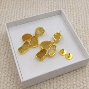 May include: Gold-plated earrings featuring irregular, amber-colored gemstones. The earrings have a cluster design with multiple stones and are accompanied by two gold-colored earring posts. The jewelry is presented in a white box.
