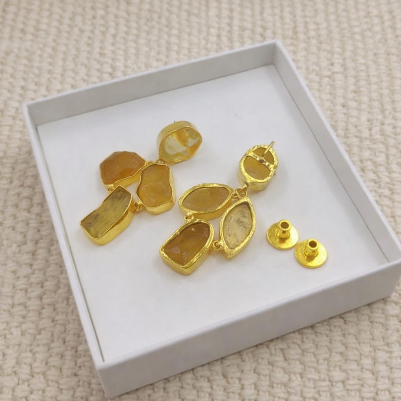 May include: Gold-plated earrings featuring irregular, amber-colored gemstones. The earrings have a cluster design with multiple stones and are accompanied by two gold-colored earring posts. The jewelry is presented in a white box.