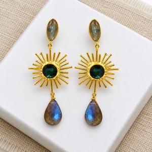 Puede incluir: Un par de pendientes colgantes dorados con un dise&ntilde;o de rayos de sol y una piedra circular verde oscuro en el centro. Cada pendiente tiene una piedra en forma de marquesa en la parte superior y una piedra azul iridiscente en forma de gota.