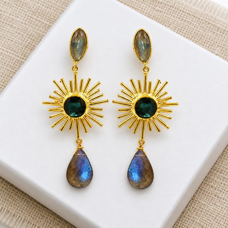 Puede incluir: Un par de pendientes colgantes dorados con un dise&ntilde;o de rayos de sol y una piedra circular verde oscuro en el centro. Cada pendiente tiene una piedra en forma de marquesa en la parte superior y una piedra azul iridiscente en forma de gota.