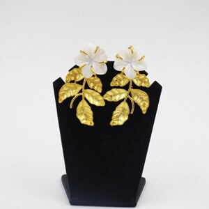 May include: A pair of floral earrings displayed on a black stand. Each earring features a white flower with gold-colored accents and a stem with gold-colored leaves. The earrings are set against a white background.