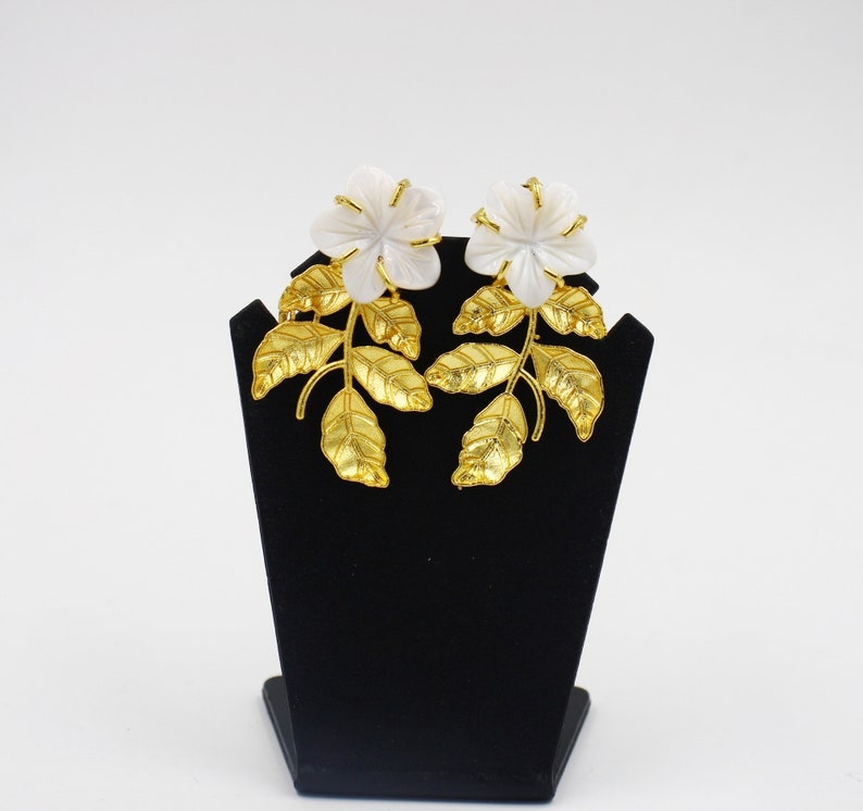 May include: A pair of floral earrings displayed on a black stand. Each earring features a white flower with gold-colored accents and a stem with gold-colored leaves. The earrings are set against a white background.